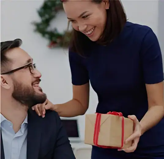 Woman giving a wrapped gift to a smiling man in an office setting, celebrating a special occasion with a focus on knitting and sock yarn.